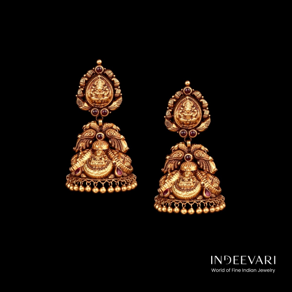 Elegant Lakshmi Nakshi Jhumkis displaying finely carved central goddess Lakshmi motif surrounded by intricate floral and peacock design elements, antique gold finish, delicate ball hangings at base, and subtle ruby stone accents