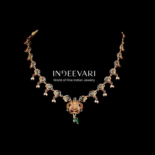 Radiant Devi Emerald Necklace featuring intricately sculpted Devi goddess pendant centerpiece, connected chain of emerald-accented peacock motifs throughout, delicate pearl dangler accents, vibrant emerald gemstone settings, antique gold finish