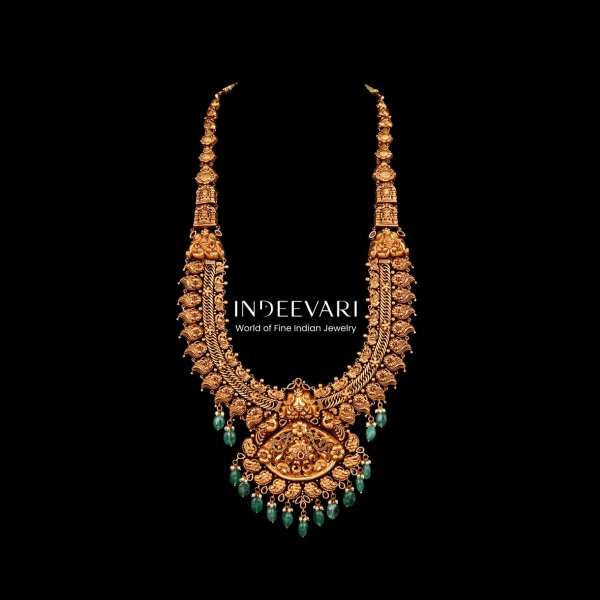 Luxurious Peacock Nakshi Emerald Haram displaying grand pendant with detailed peacock and floral carved motifs, intricate nakshi work throughout in antique gold, clusters of green emerald gemstone drops positioned at intervals, golden bead chain elements,